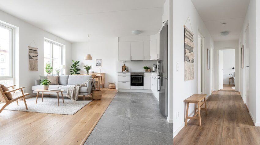 Ljust modernt vardagsrum och kök i öppen planlösning med vita väggar, trä- och klinkergolv, grå soffa, trämöbler, växter och en hall som leder till andra rum. Stora fönster släpper in naturligt ljus.